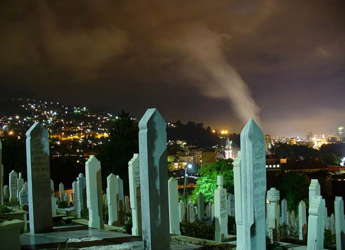A hillside graveyard of white headstones at night, the lights of a city spread out below